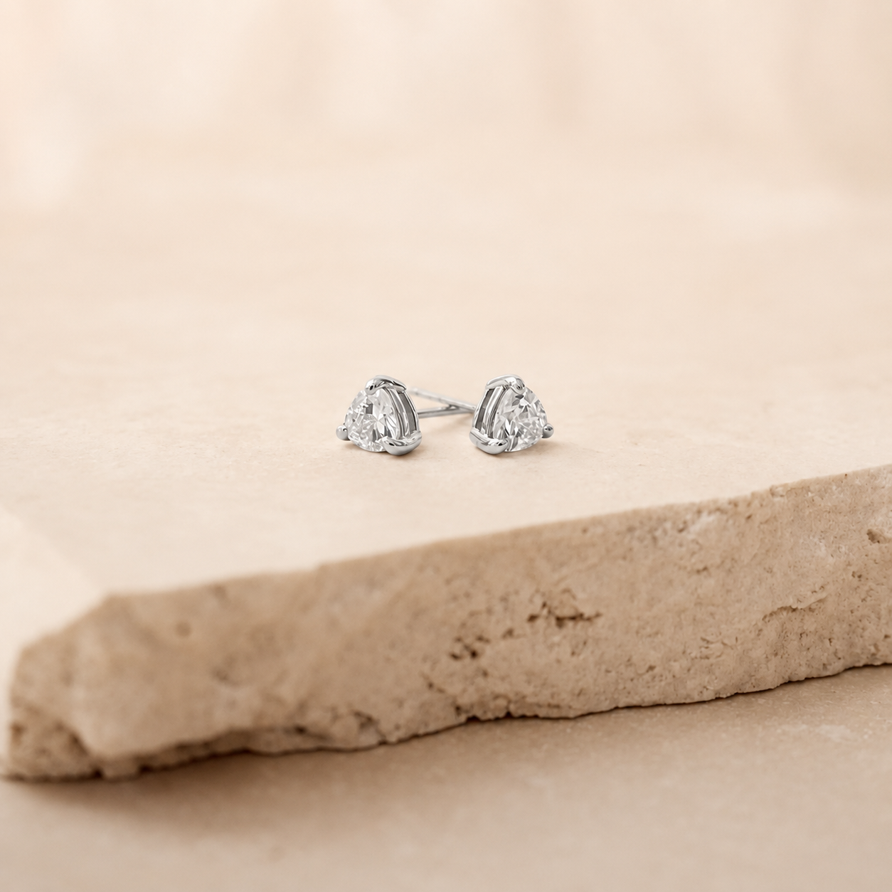 A pair of silver stud earrings with triangular clear gemstones, resting on a textured beige stone slab against a soft beige background.