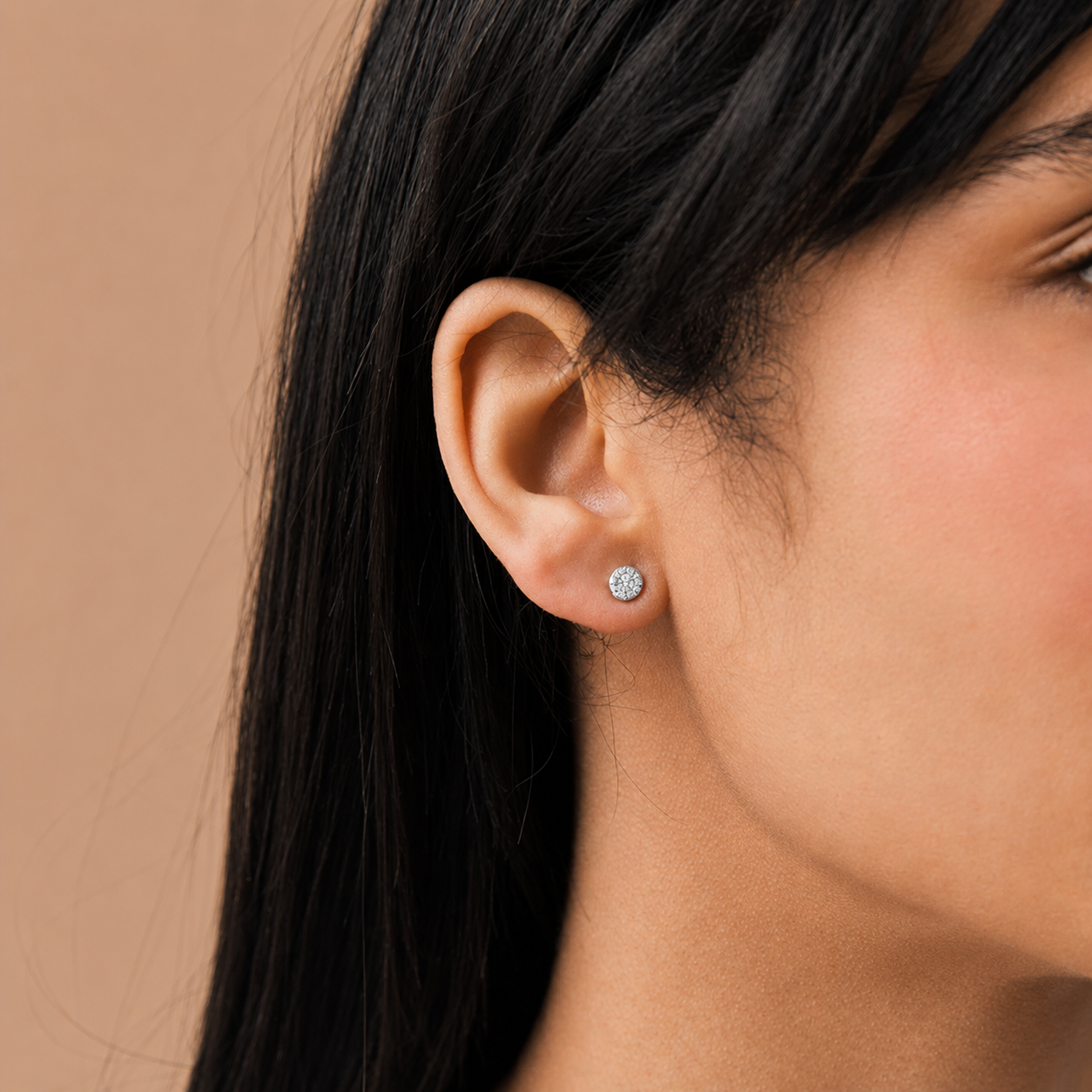 A close-up of a woman's ear wearing a small round stud earring with a clear gemstone in a silver-toned setting, against a warm beige background.