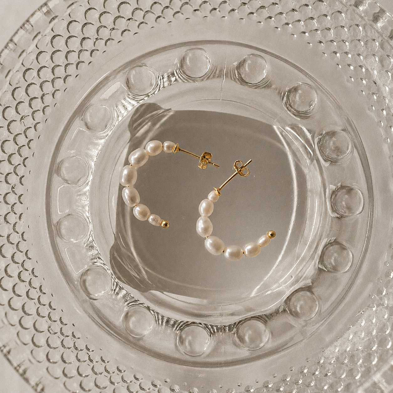A pair of gold hoop earrings with small white pearl beads, displayed on a clear glass dish over a textured white surface.