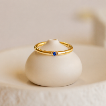 Gold ring with a blue gemstone on a white ceramic stand against a beige background