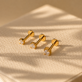 Three gold bar-shaped earrings with clear gemstones on a beige fabric background