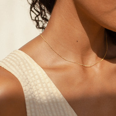 A close-up image of a person wearing a dainty satellite necklace with metal bead detailing, in gold color.