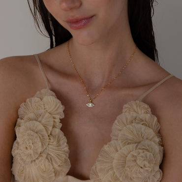A person wearing a beige top showcasing a gold-colored evil eye protection necklace with a pave charm.