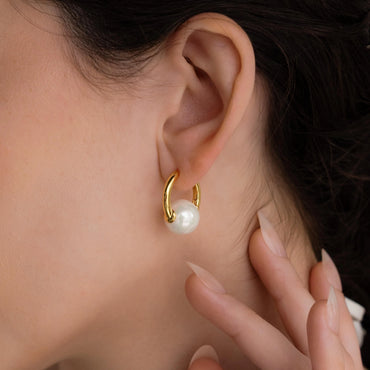 A close-up image of a woman's ear wearing a gold-colored hoop earring with a round pearl charm.