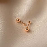 Two rose gold barbell stud earrings with spherical beads on a textured beige fabric background.