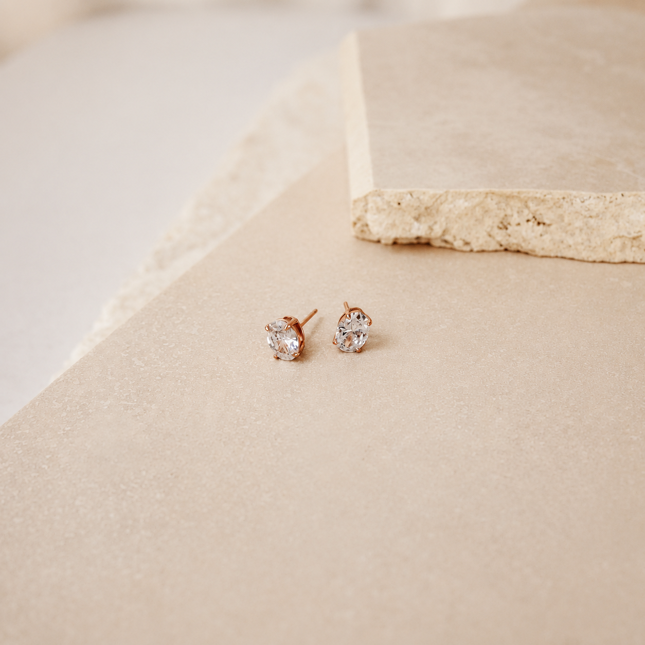 A pair of rose gold stud earrings with clear gemstones, resting on a textured beige stone slab.