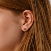 A close-up of a person's ear wearing a small round gold stud earring, with brown hair framing the side of the face.