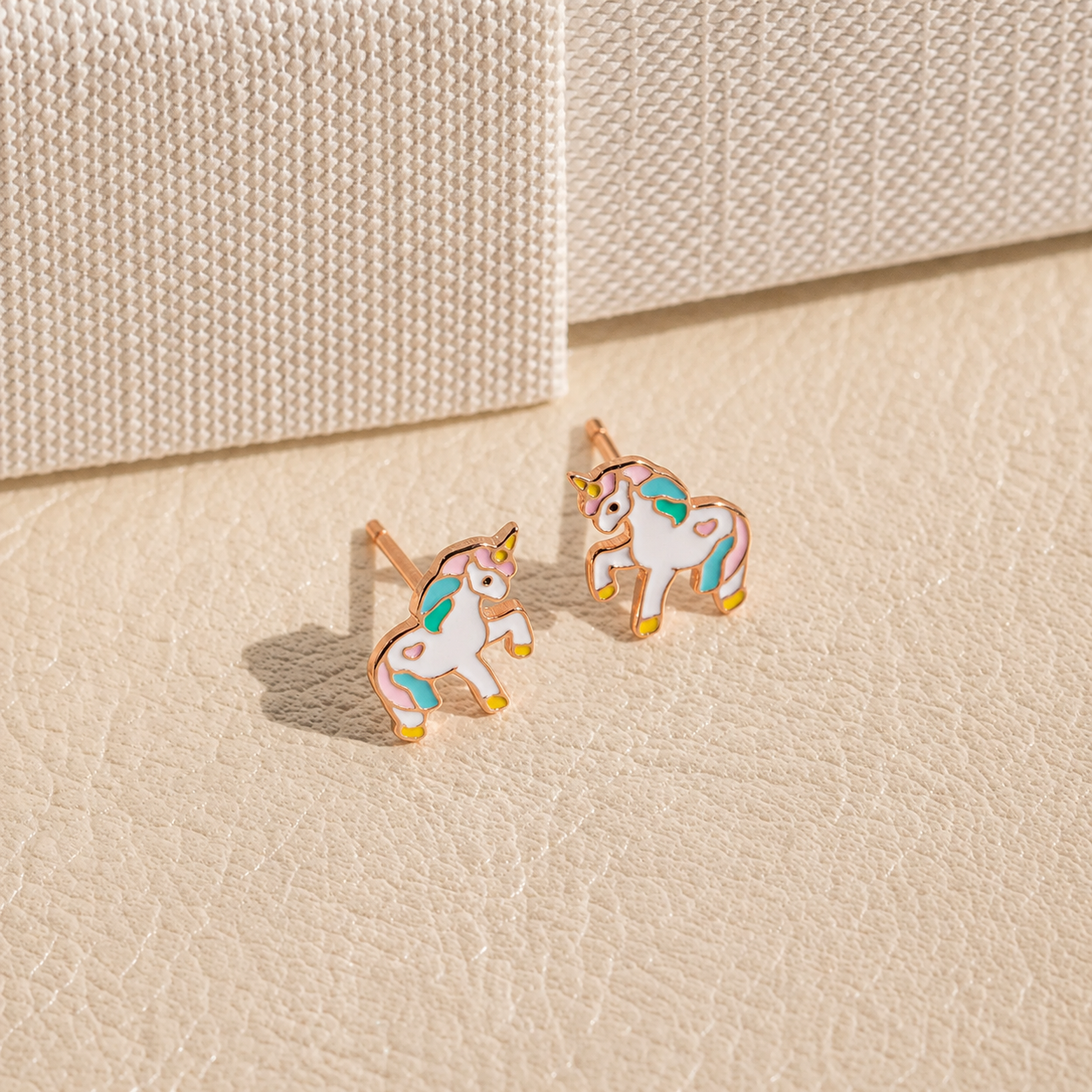 A pair of small unicorn stud earrings with white bodies, pastel pink, teal, and yellow accents, outlined in gold-tone metal, displayed on a textured beige surface.