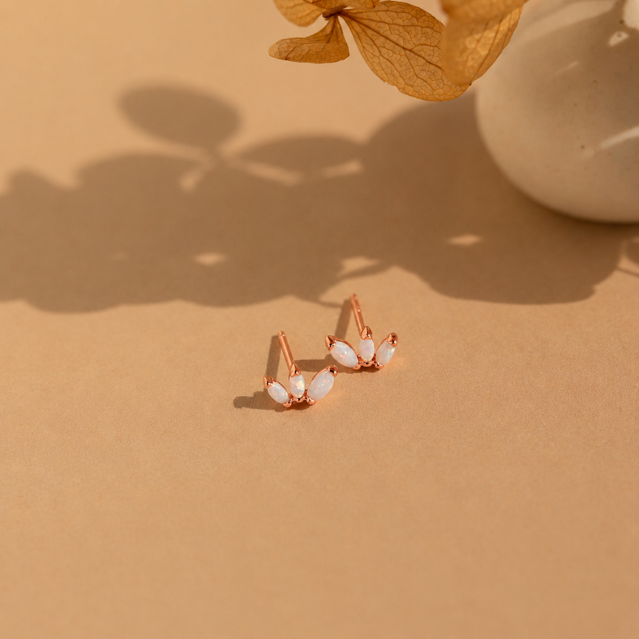 Rose gold earrings with white stones on a beige surface with a leaf and vase in the background