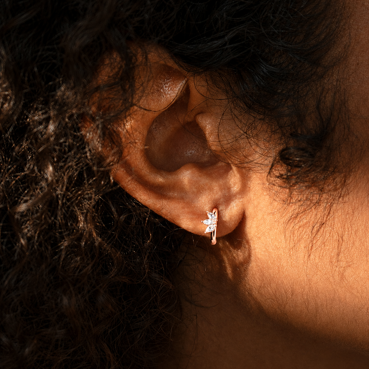 A close-up of a person's ear wearing a small silver earring with a white flower and a tiny red accent, set against dark curly hair under warm, dramatic lighting.