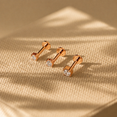 Rose gold barbell-shaped earrings with clear gemstones on a beige fabric background