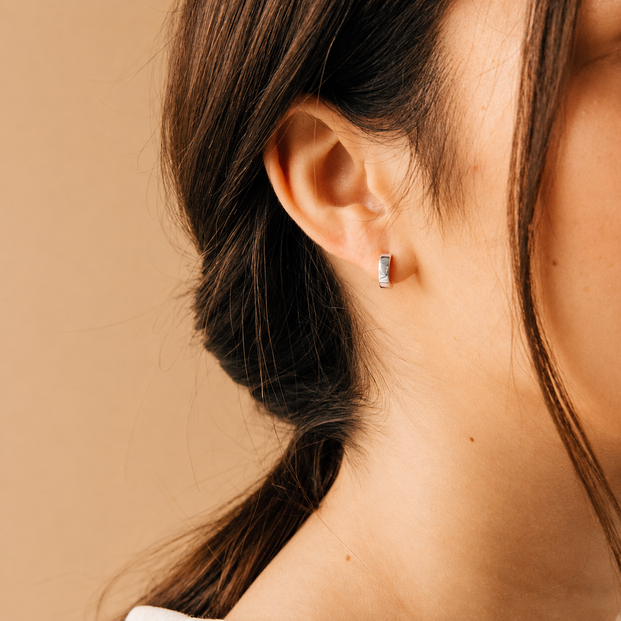 A close-up of a woman's ear wearing a small silver huggie hoop earring with clear stones, set against a warm beige background.