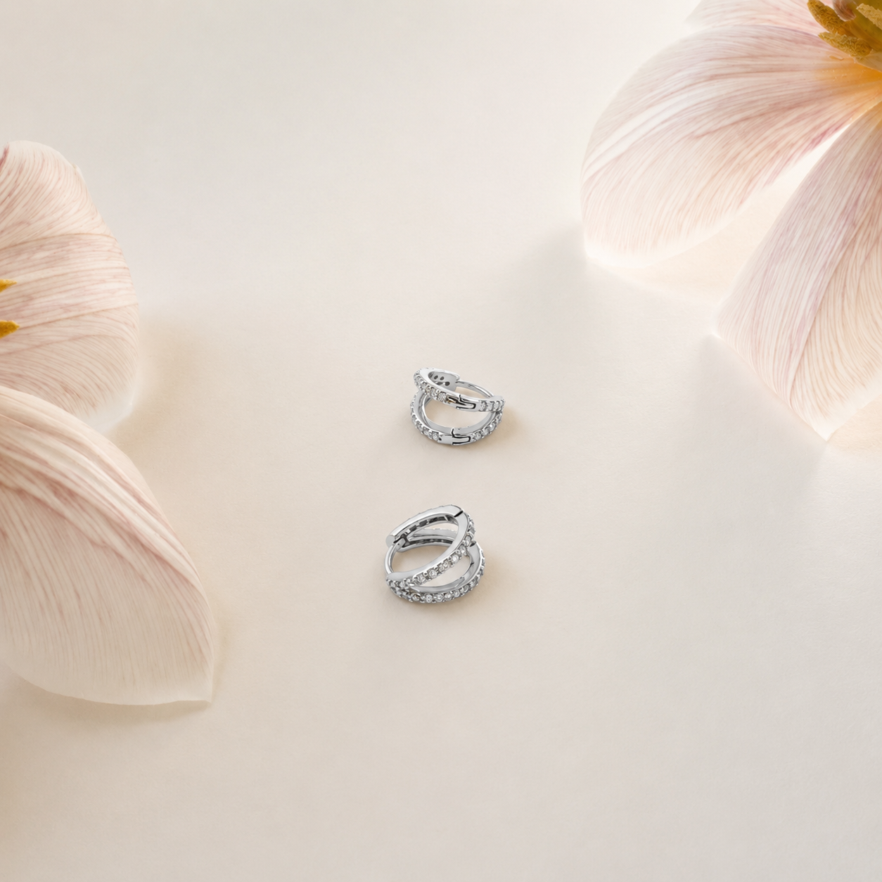 A pair of silver-toned huggie hoop earrings with textured, snake-like ridges, displayed on a warm cream surface with soft pink flower petals in the corners.