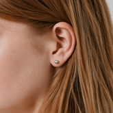 A close-up of a person's ear wearing a small round silver stud earring, with brown hair framing the side of the face.