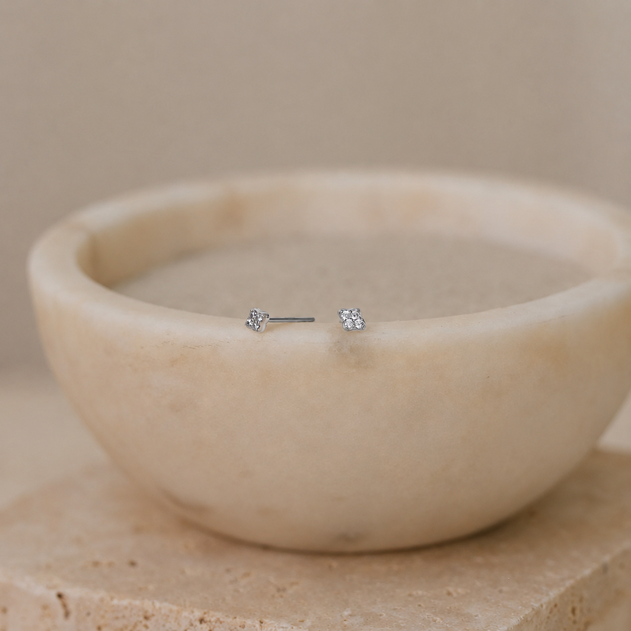 A pair of small silver stud earrings with clear stones, displayed on the rim of a round beige stone bowl.