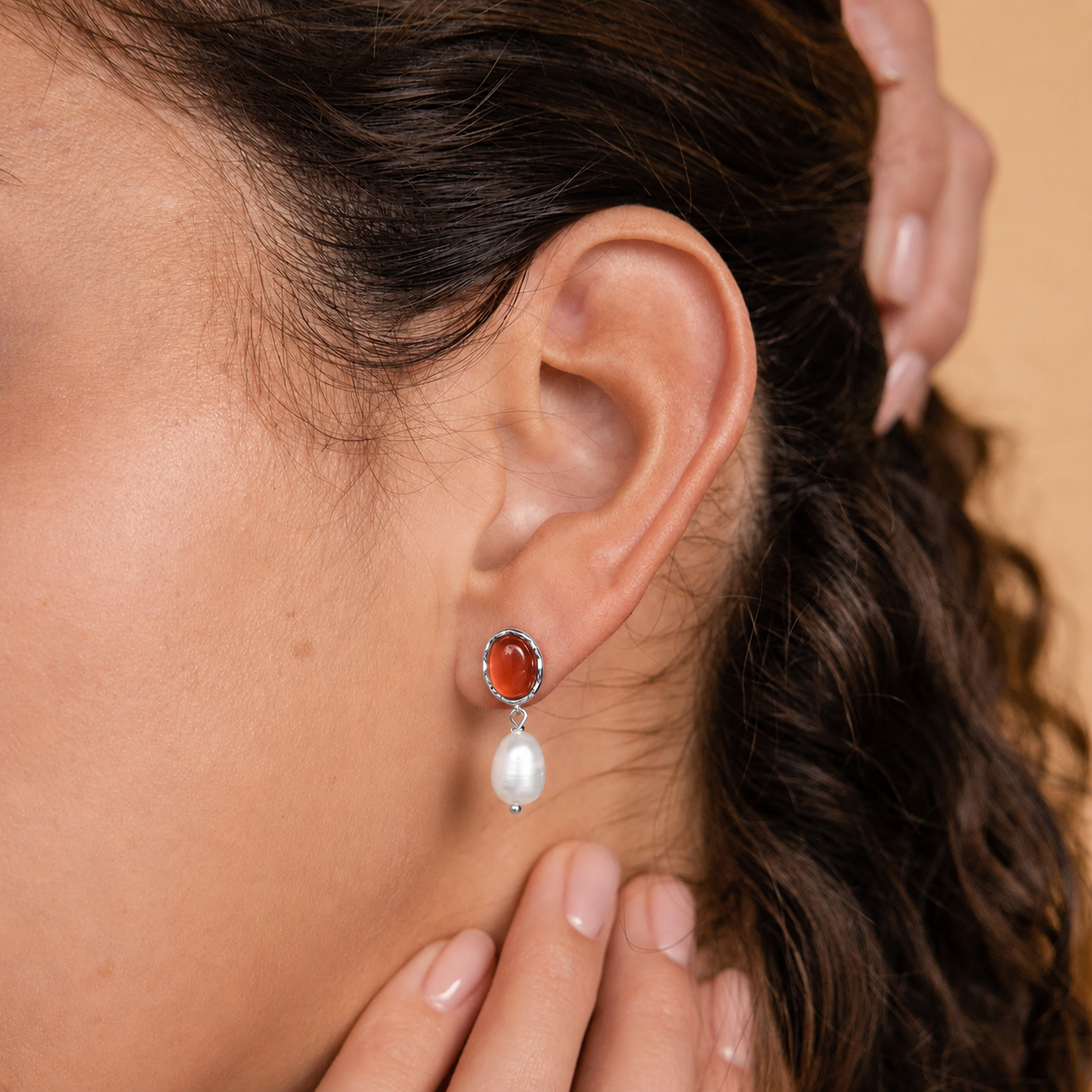 Close-up of an ear wearing a silver earring with a red gemstone and pearl drop, with a hand adjusting hair.