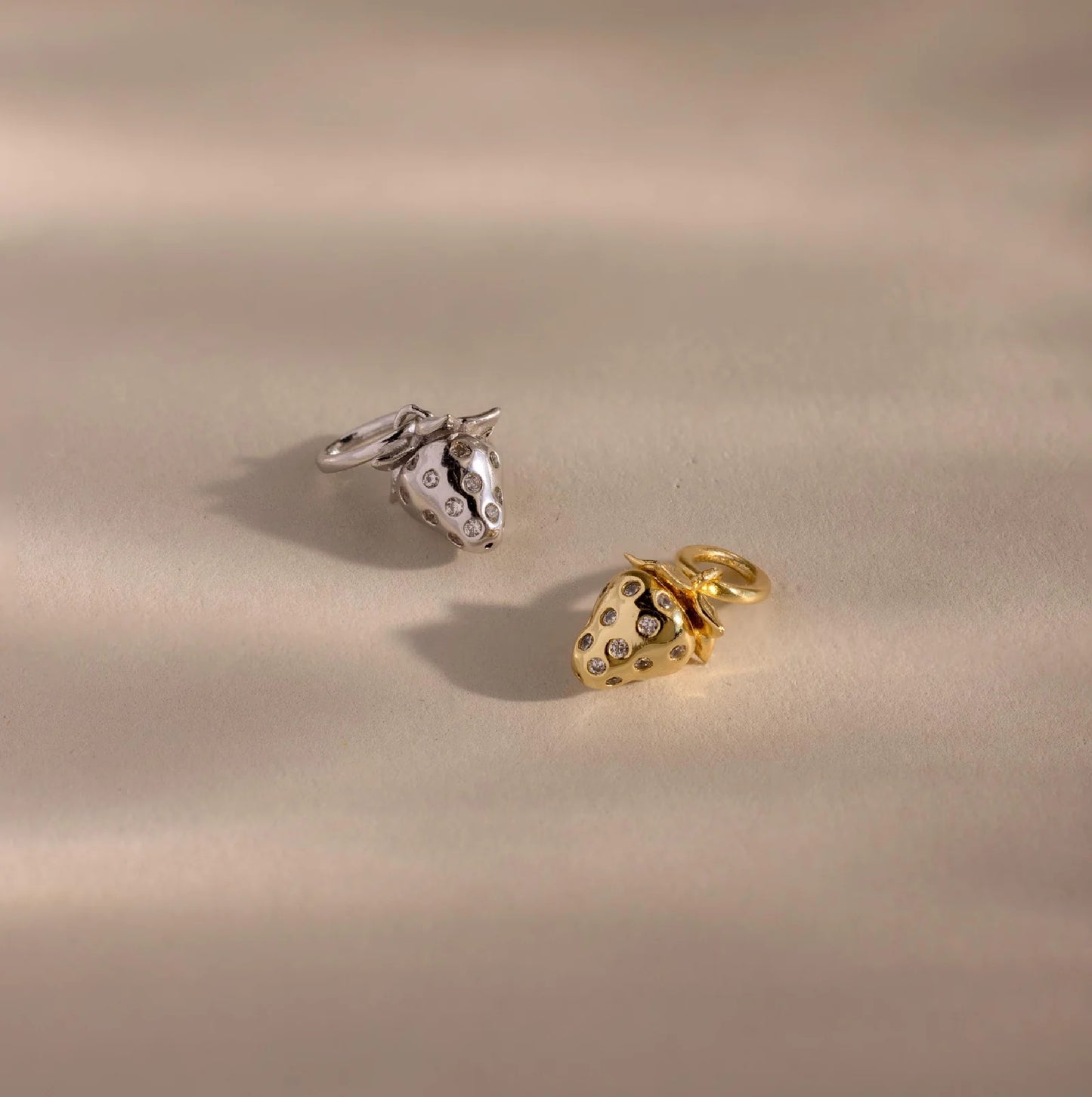 Two strawberry-shaped charms, one gold and one silver, on a beige background.