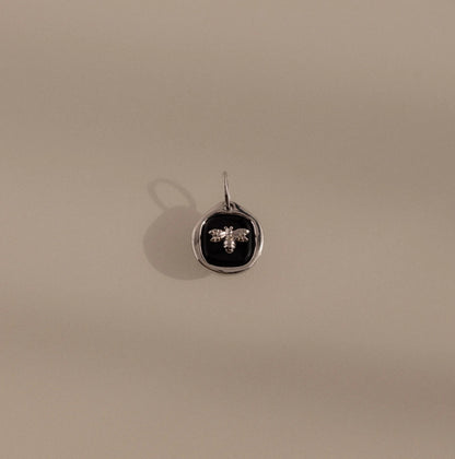 Silver pendant with a black circle and bee design on a beige background