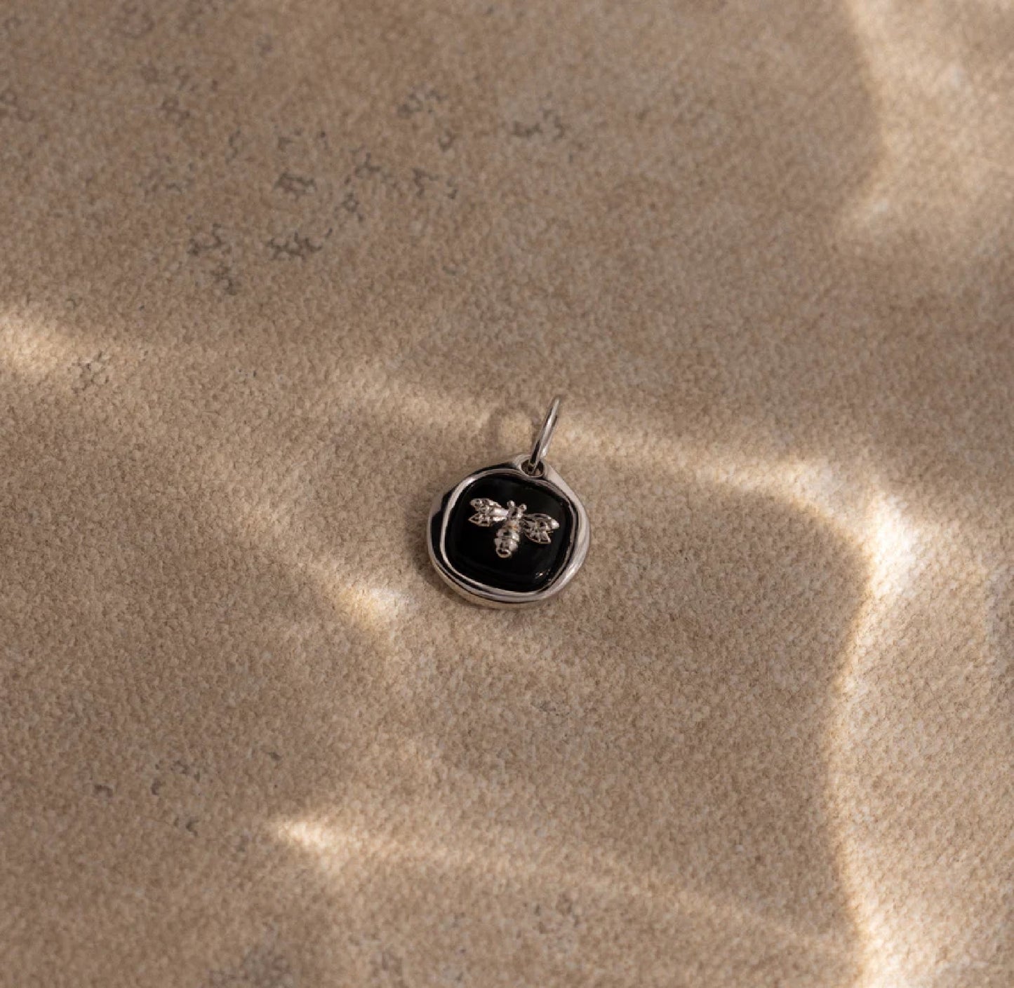 Silver pendant with a black stone and bee design on a textured beige surface