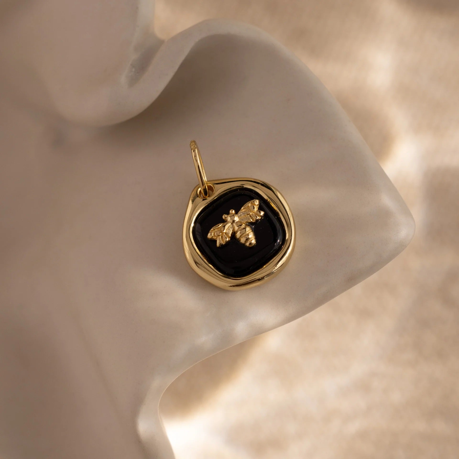 Gold pendant with a black stone and bee design on a soft beige background