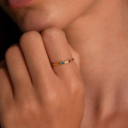 Close-up of a hand wearing a gold ring with colored stones, touching the chin.