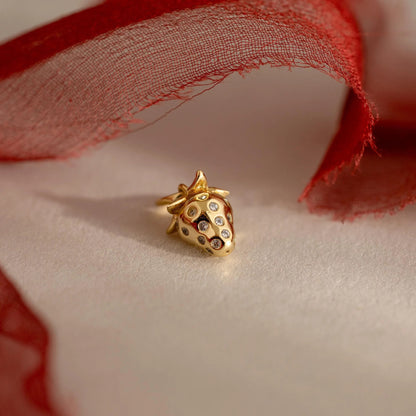 Gold strawberry-shaped jewelry piece with embedded stones on a soft fabric background