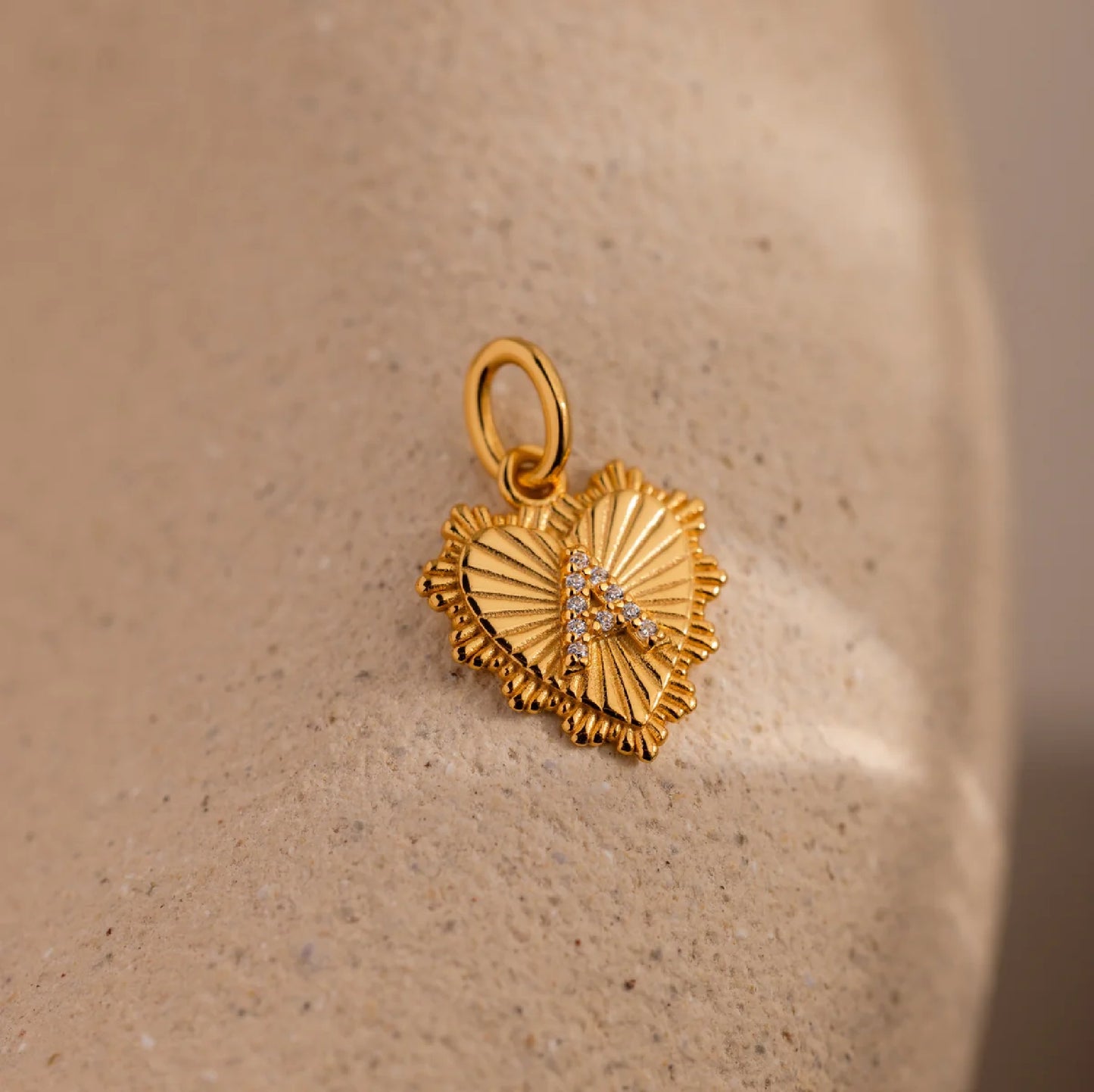 Gold heart-shaped pendant with letter 'A' on a beige stone surface