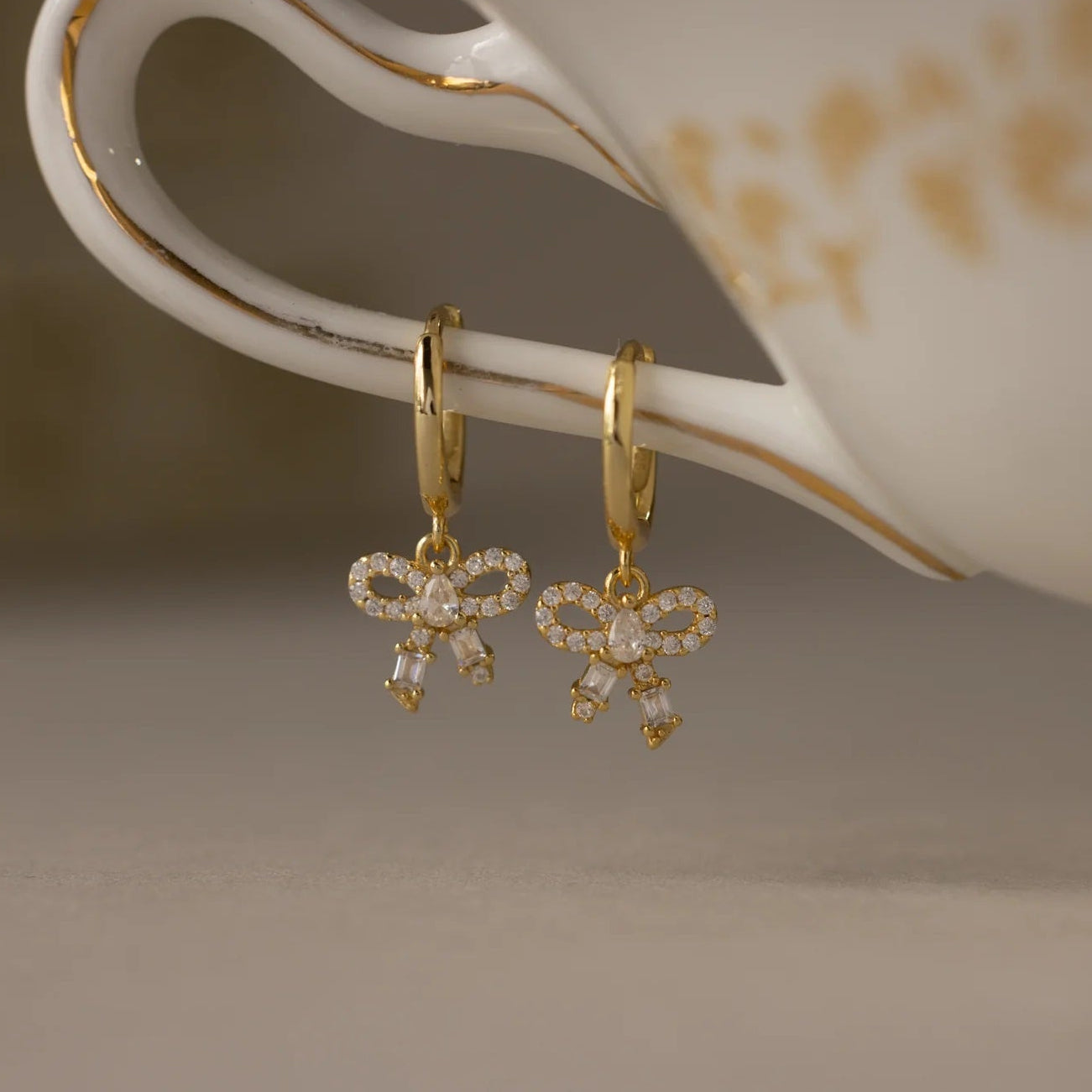Gold bow-shaped earrings hanging on a white teacup with gold floral patterns.
