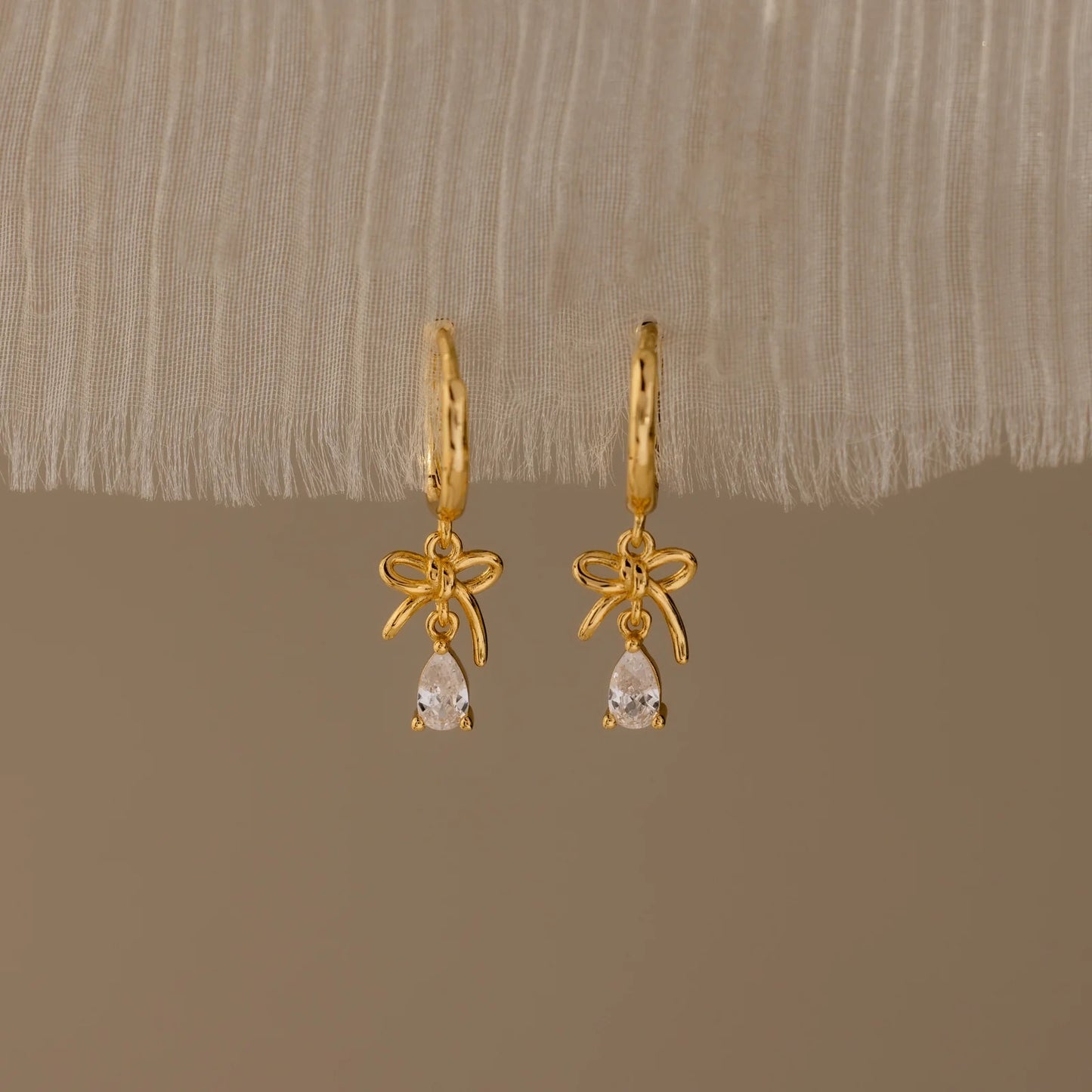 Gold earrings with bow and teardrop design on a beige fabric background