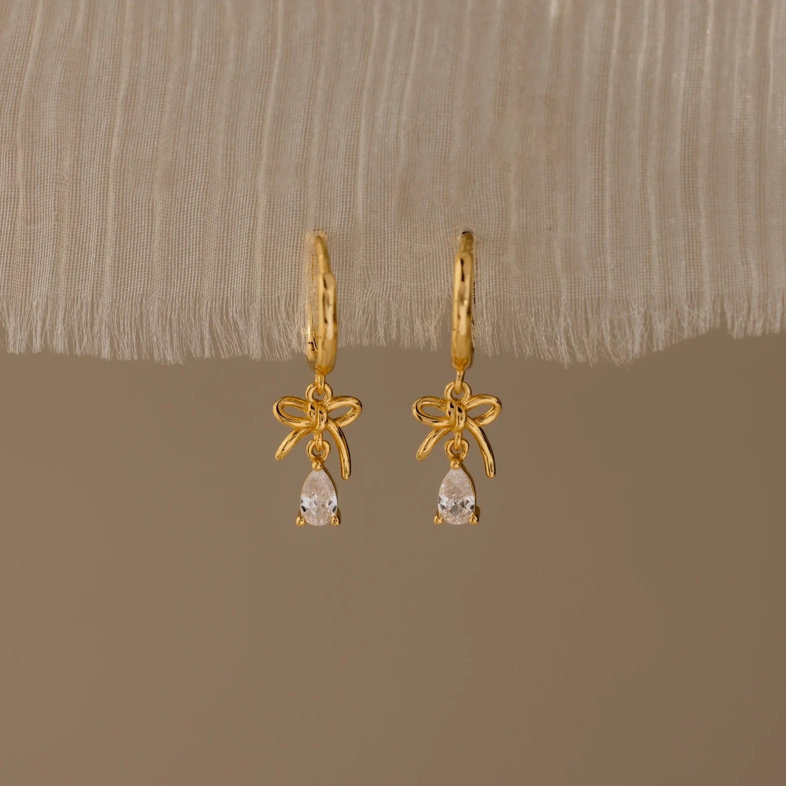 Gold earrings with bow and teardrop design on a beige fabric background