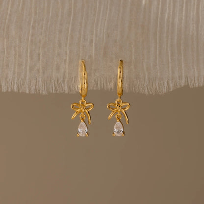 Gold earrings with bow and teardrop design on a beige fabric background