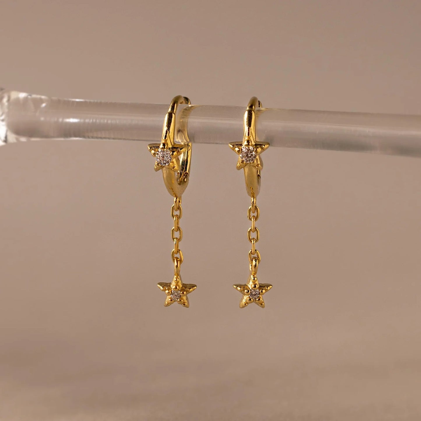 Gold hoop earrings with star-shaped pendants on a clear stand against a beige background