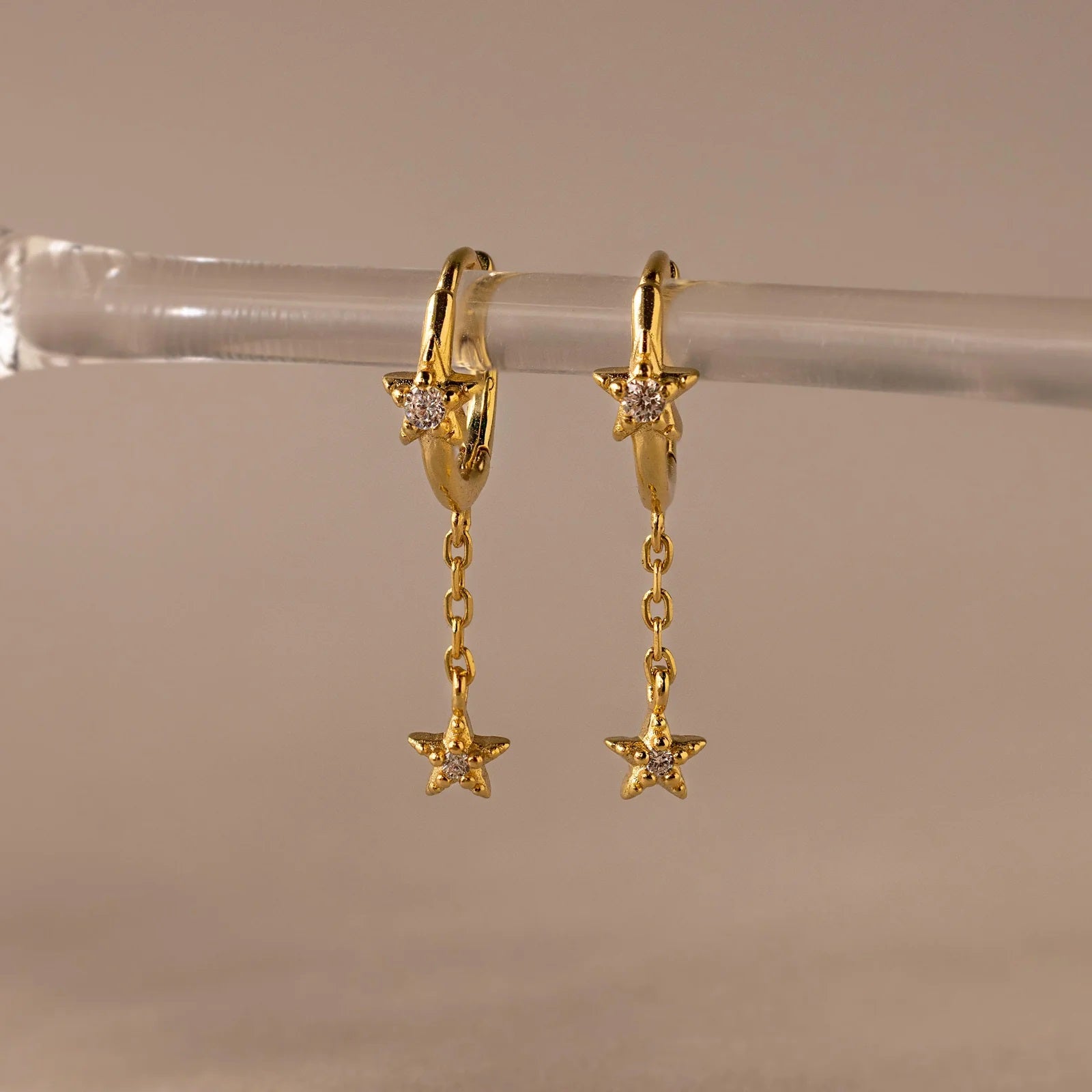 Gold hoop earrings with star-shaped pendants on a clear stand against a beige background