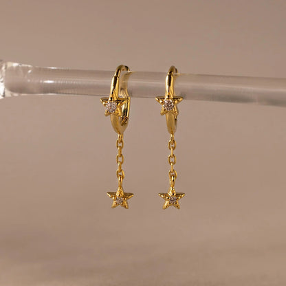 Gold hoop earrings with star-shaped pendants on a clear stand against a beige background