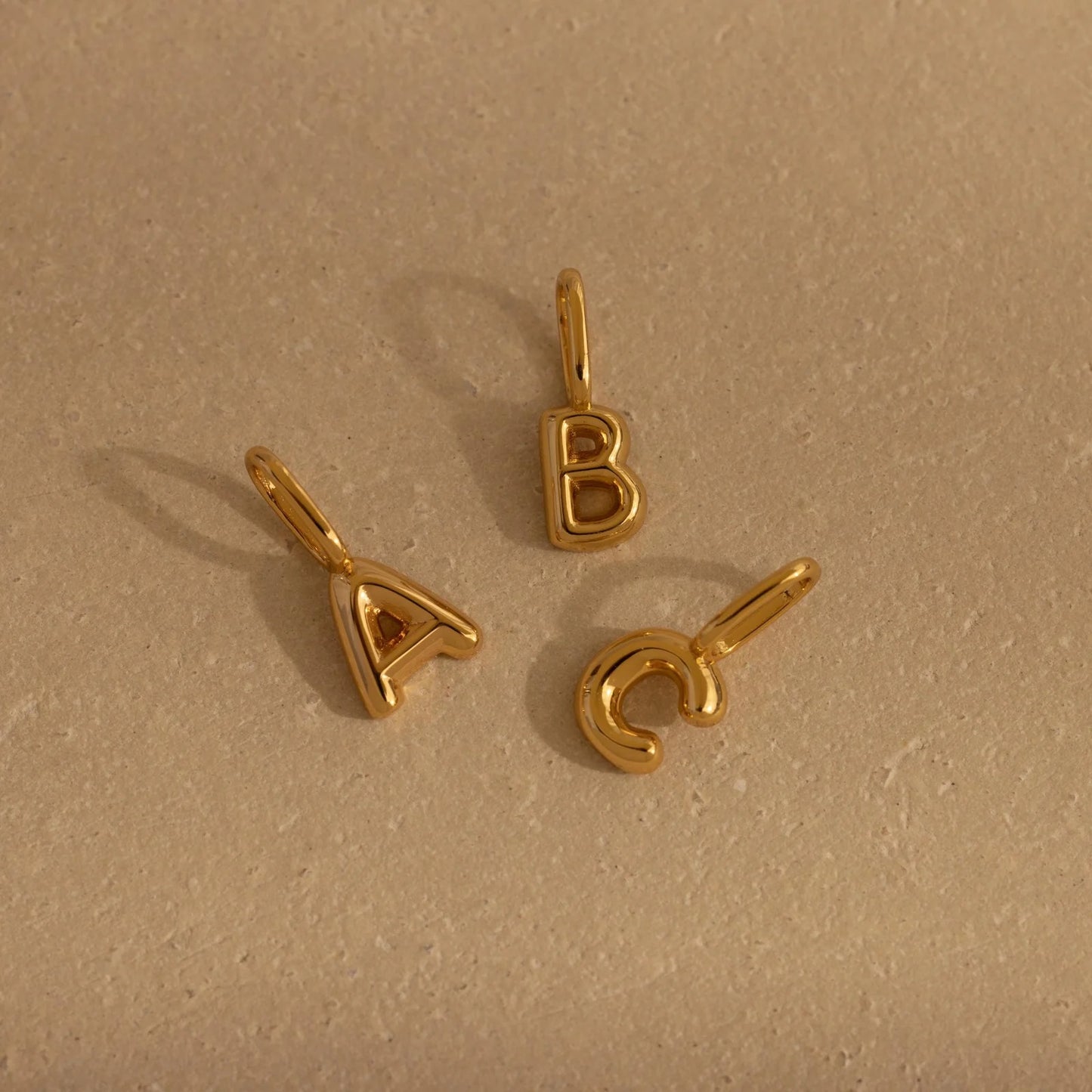 Three gold letter pendants (A, B, C) on a beige background