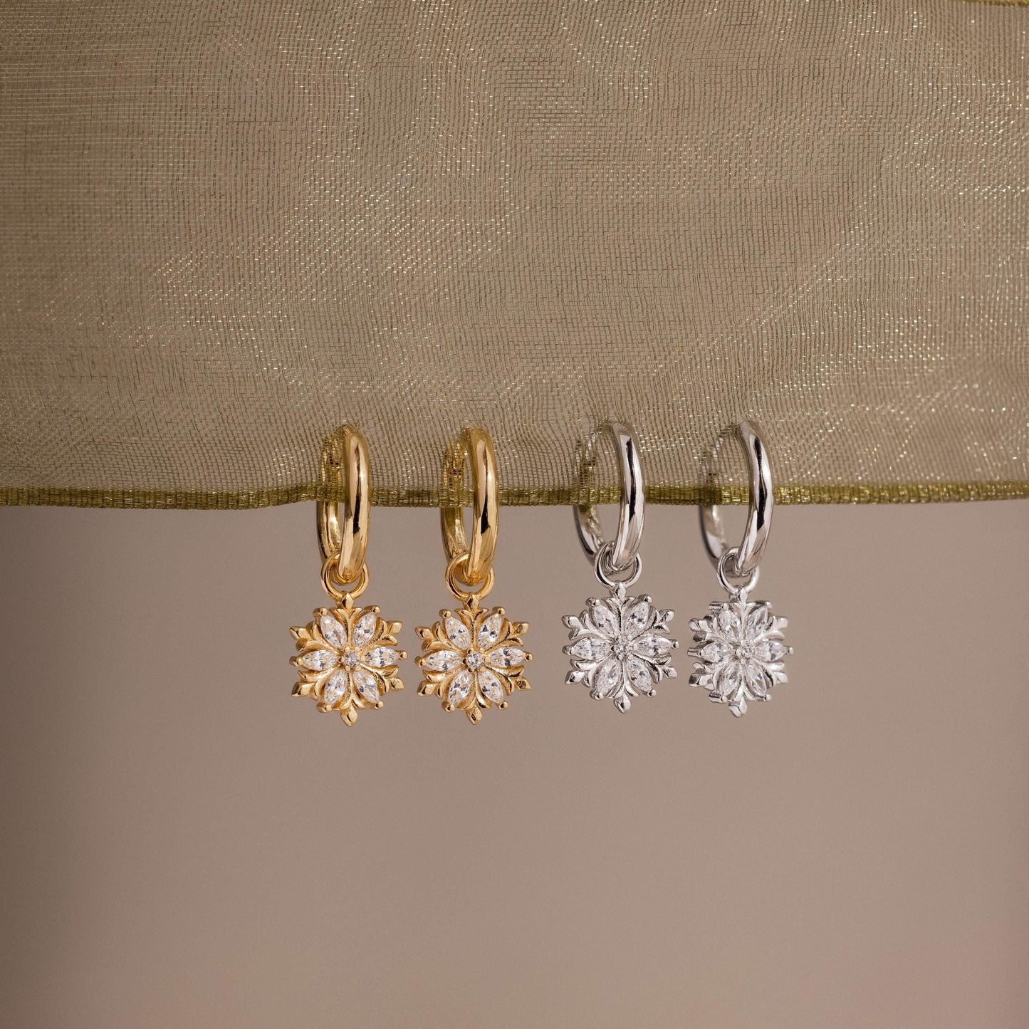 Two pairs of gold and silver hoop earrings with floral designs on a beige fabric background.