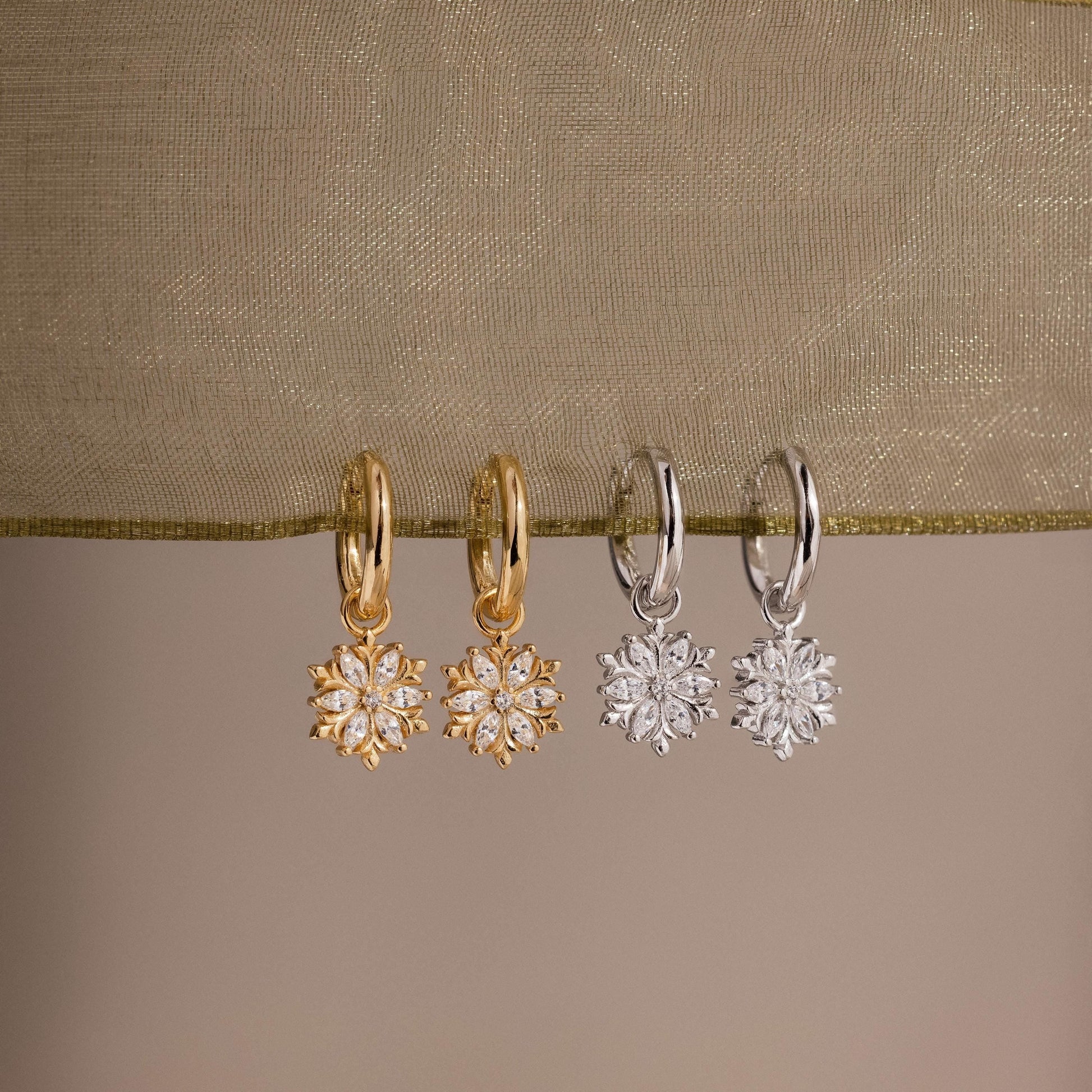 Two pairs of gold and silver hoop earrings with floral designs on a beige fabric background.