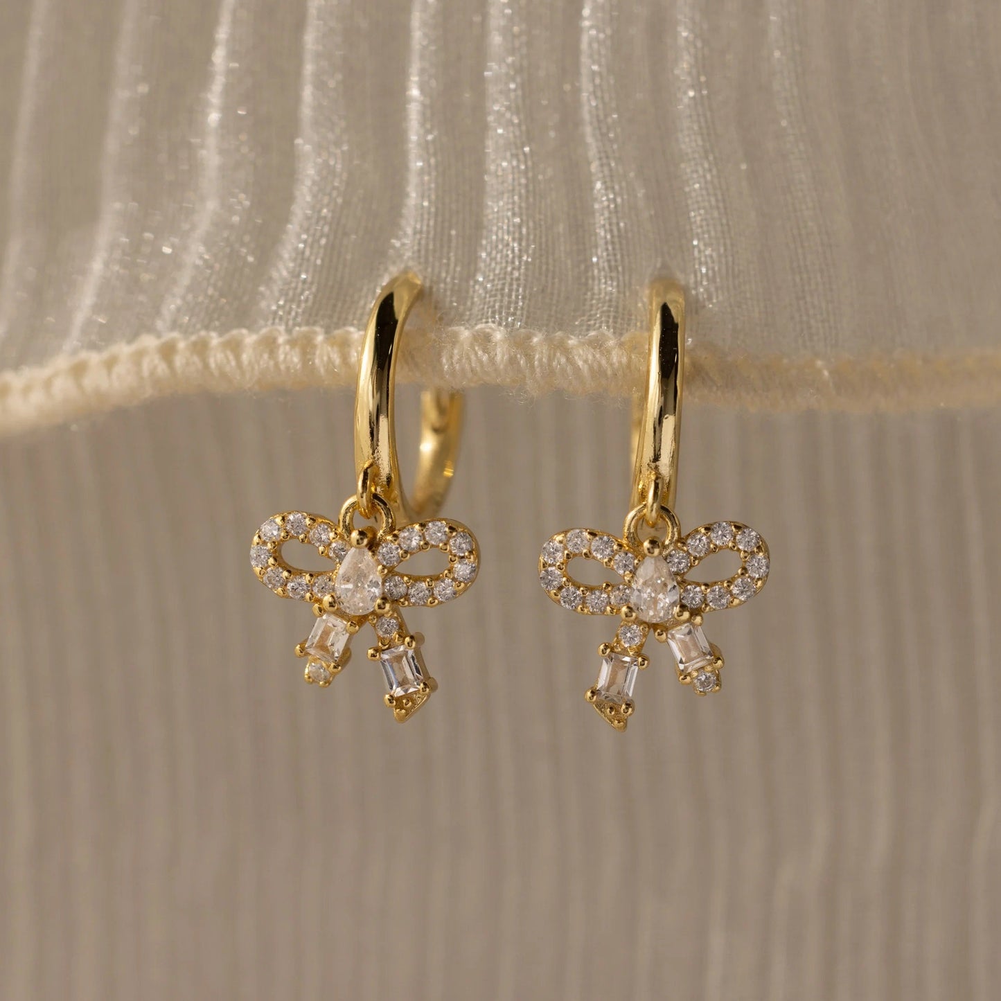 Gold bow-shaped earrings with clear stones on a beige fabric background