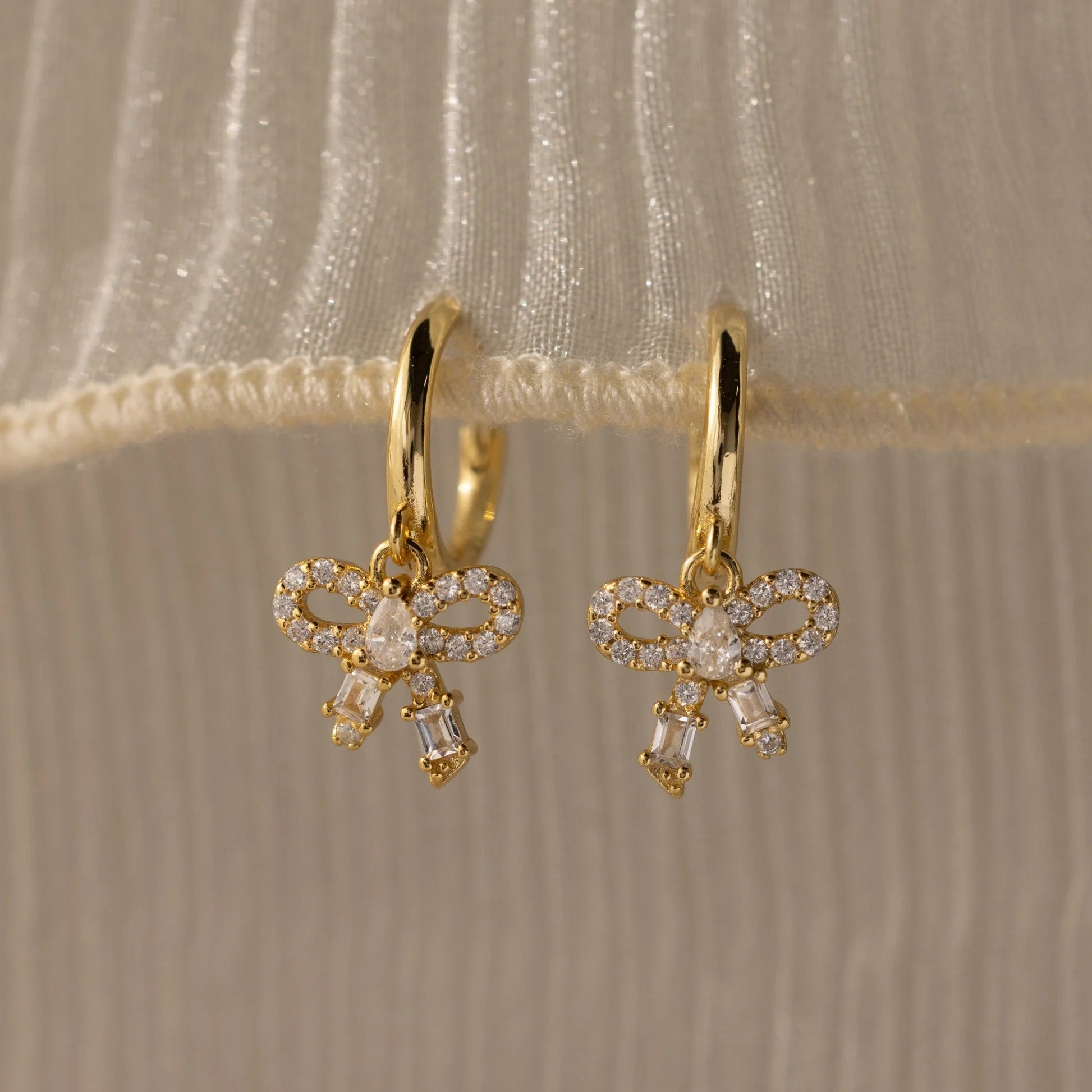 Gold bow-shaped earrings with clear stones on a beige fabric background