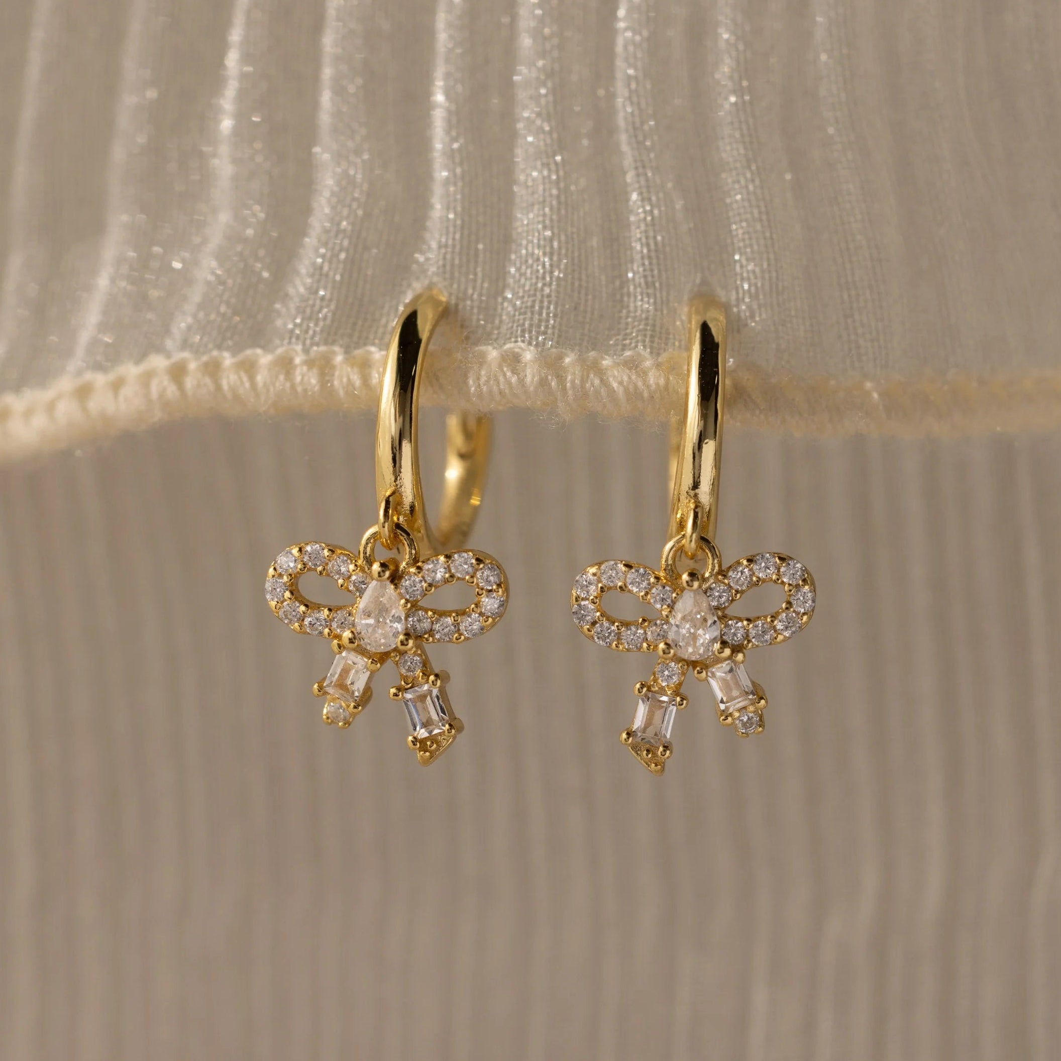 Gold bow-shaped earrings with clear stones on a beige fabric background