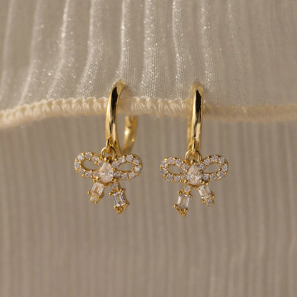 Gold bow-shaped earrings with clear stones on a beige fabric background