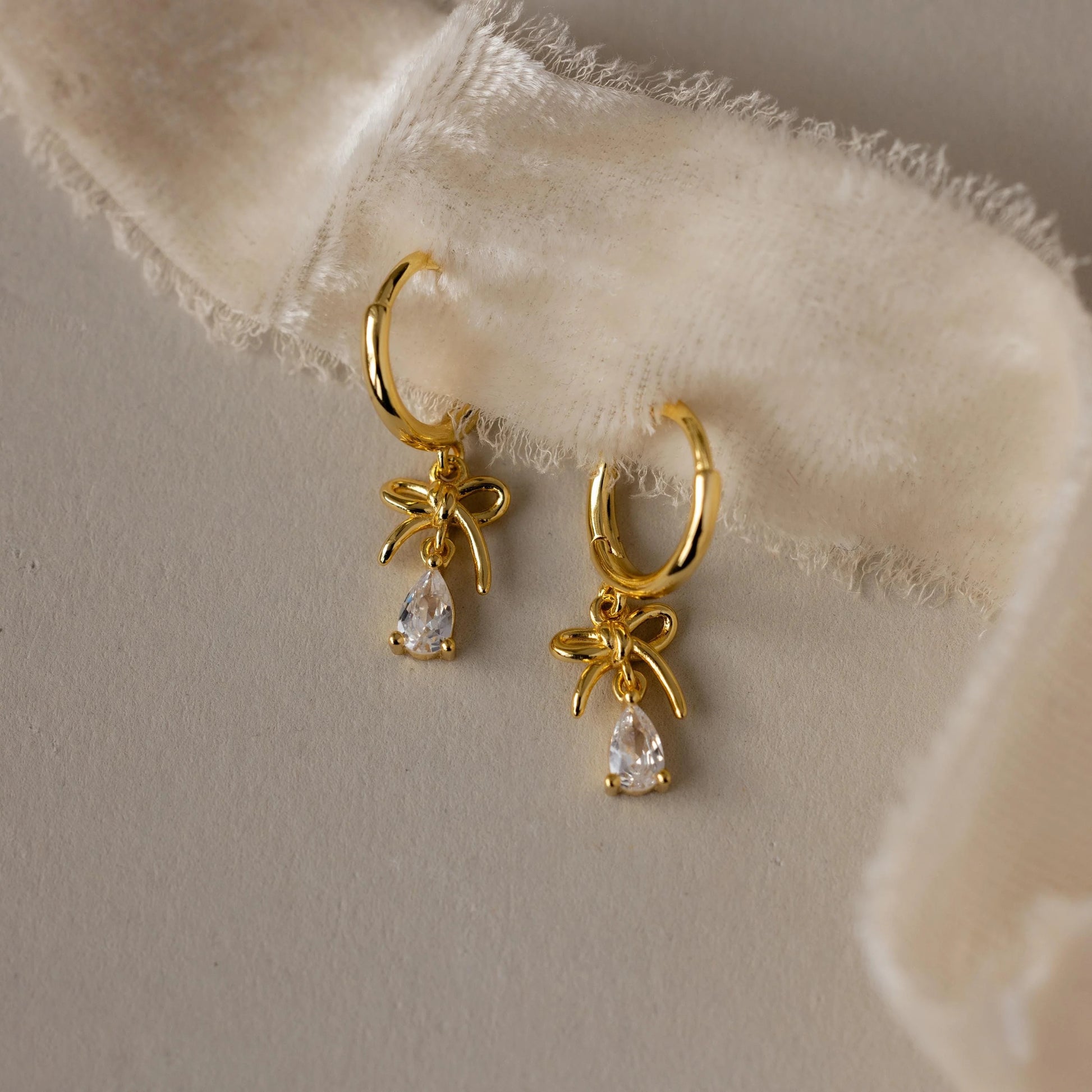 Gold hoop earrings with bow and teardrop charm on a soft fabric background