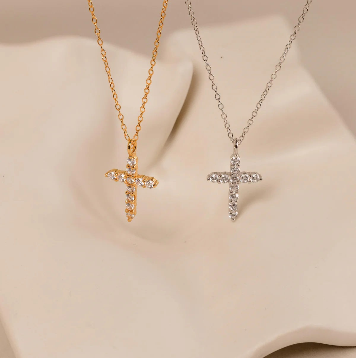 Two cross-shaped pendants on gold and silver chains against a beige background