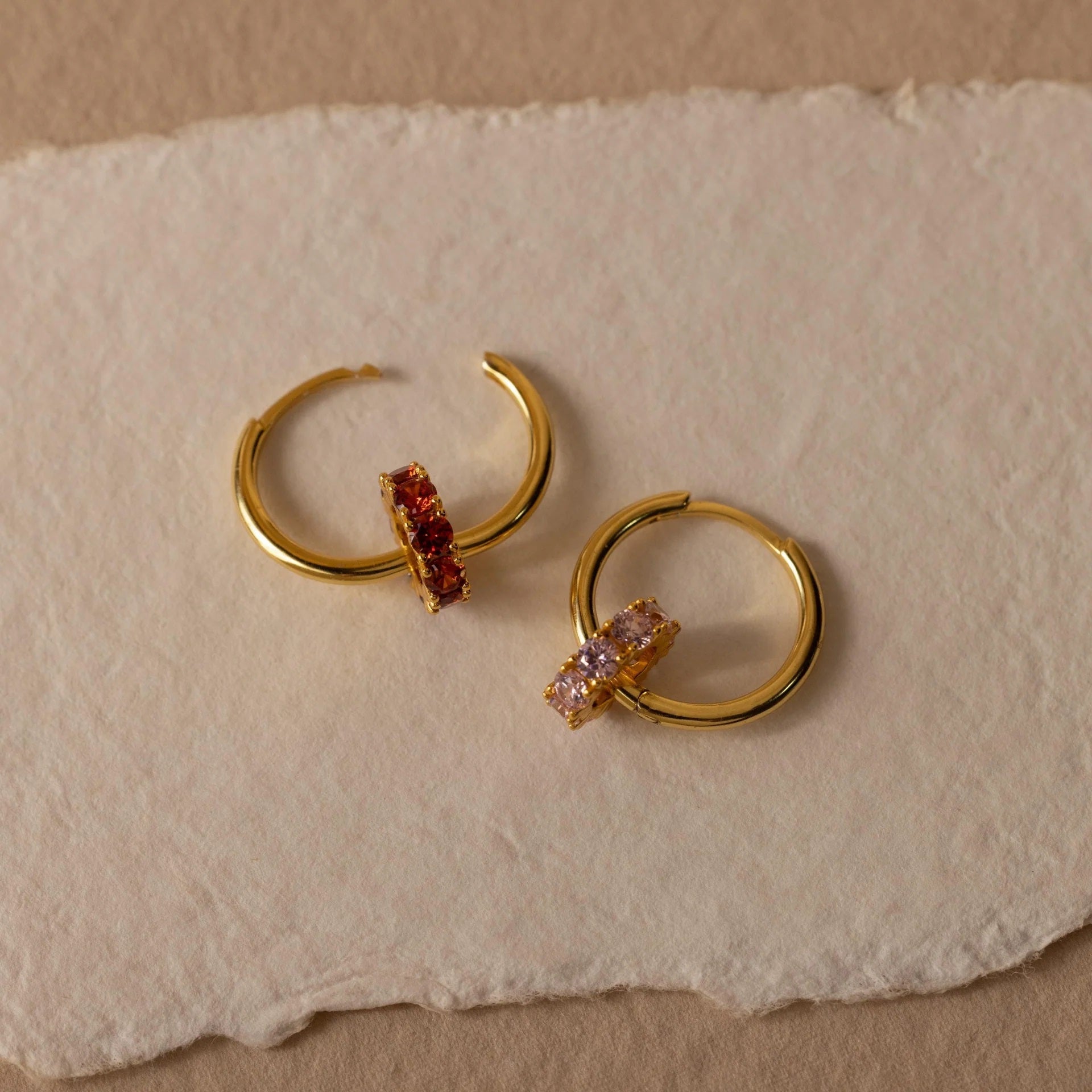Gold hoop earrings with embedded gemstones on a textured beige surface