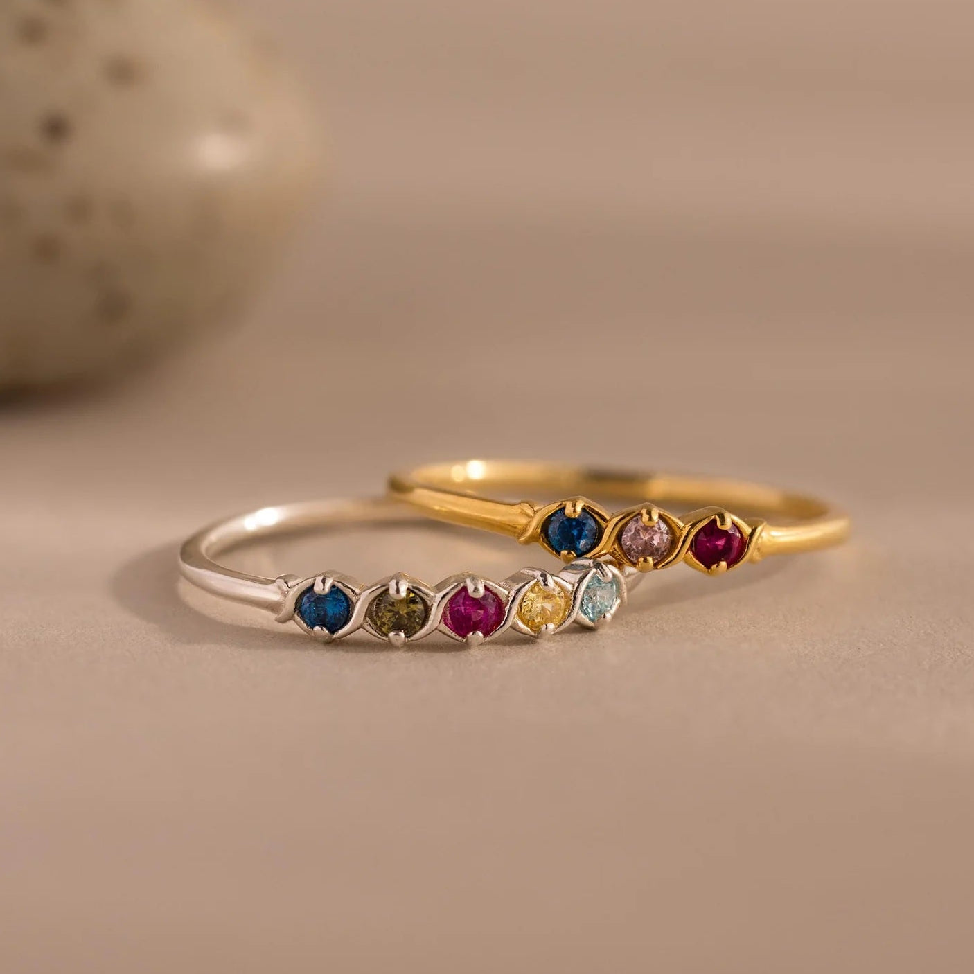 Two rings, one silver and one gold, with colorful gemstones on a beige background.