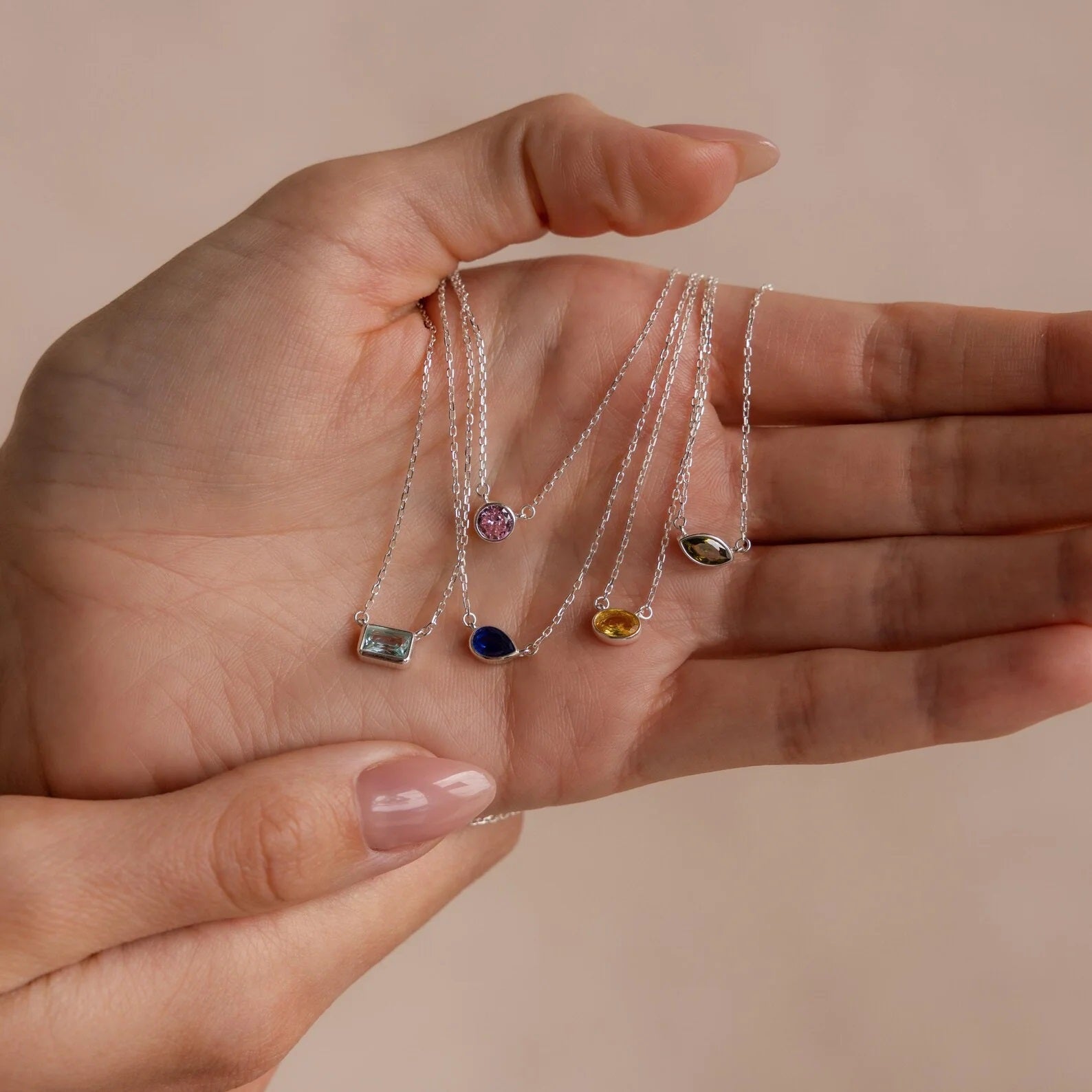 Hand holding four delicate silver necklaces with gemstone pendants against a beige background
