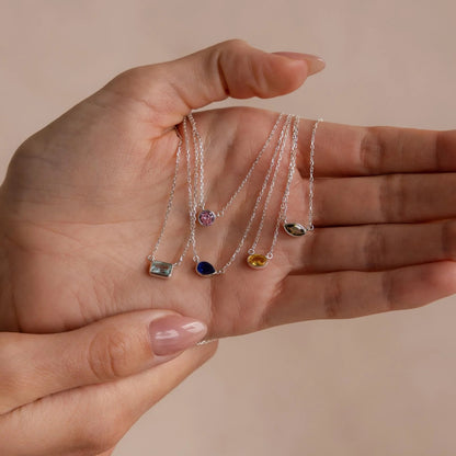 Hand holding four delicate silver necklaces with gemstone pendants against a beige background