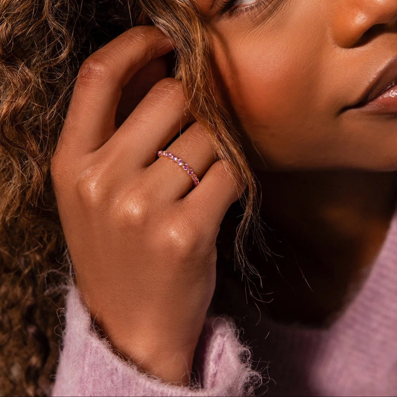 Woman with curly hair touching her face, wearing a pink sweater and ring.
