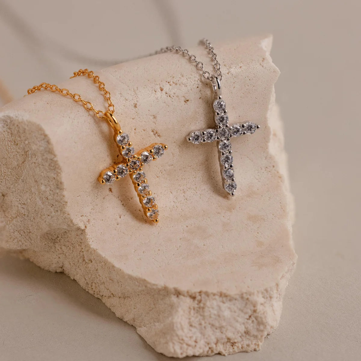 Two cross-shaped necklaces, one gold and one silver, on a textured stone surface.