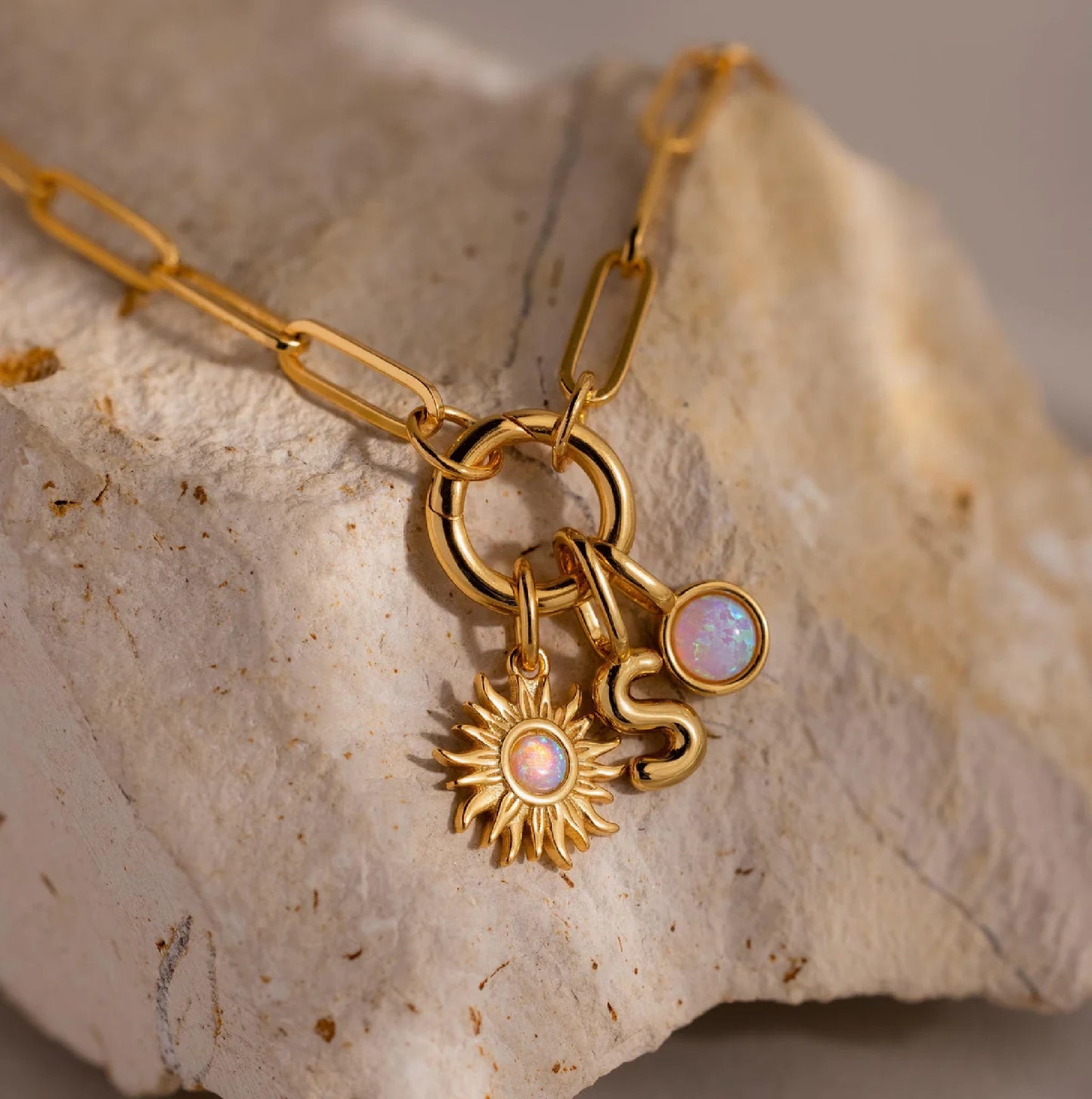 Gold necklace with sun and letter 'S' charms on a stone surface